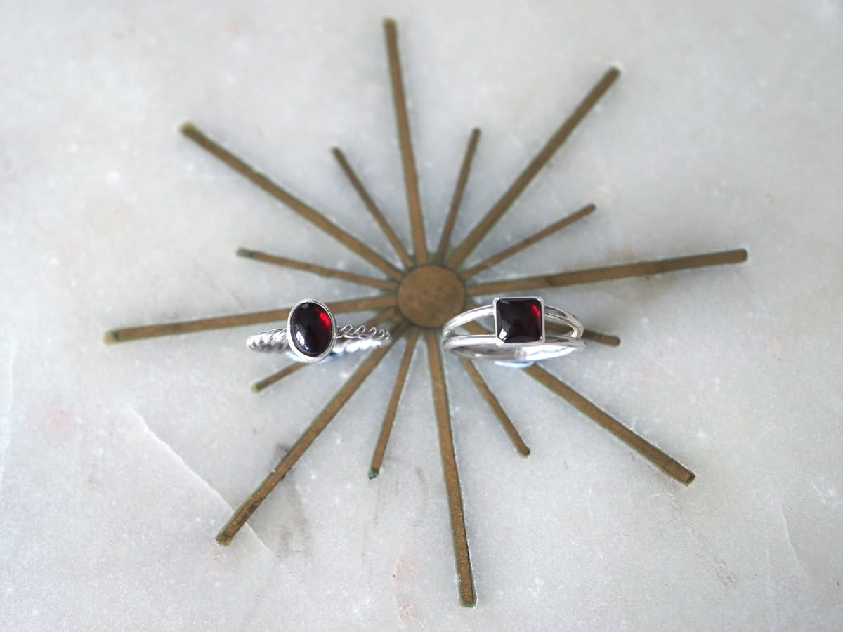 Two silver rings with garnet gemstones displayed on a brown starburst stand against a light marble background.