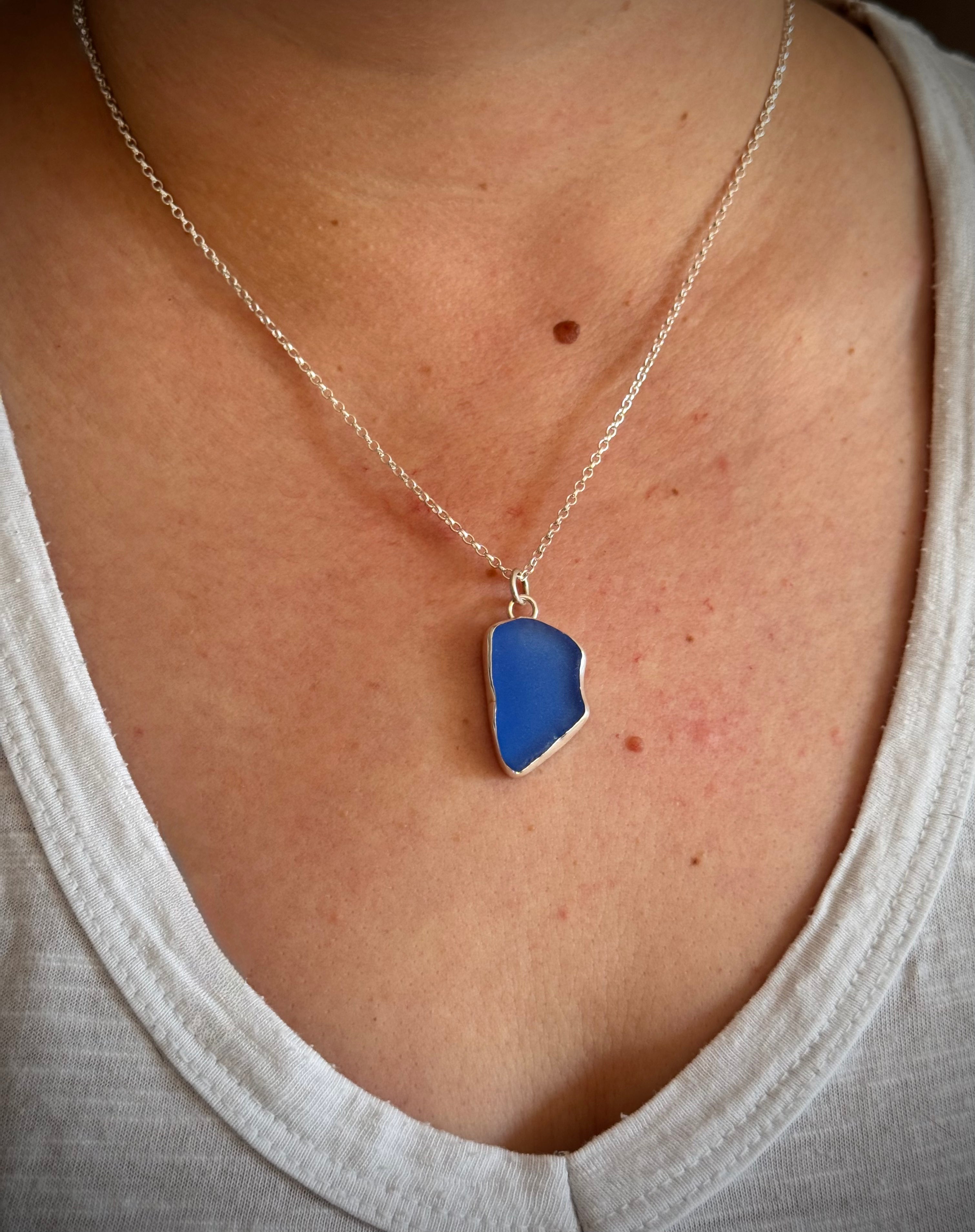 A close-up of a person wearing a silver chain necklace with a blue sea glass pendant, styled with a white v-neck top.