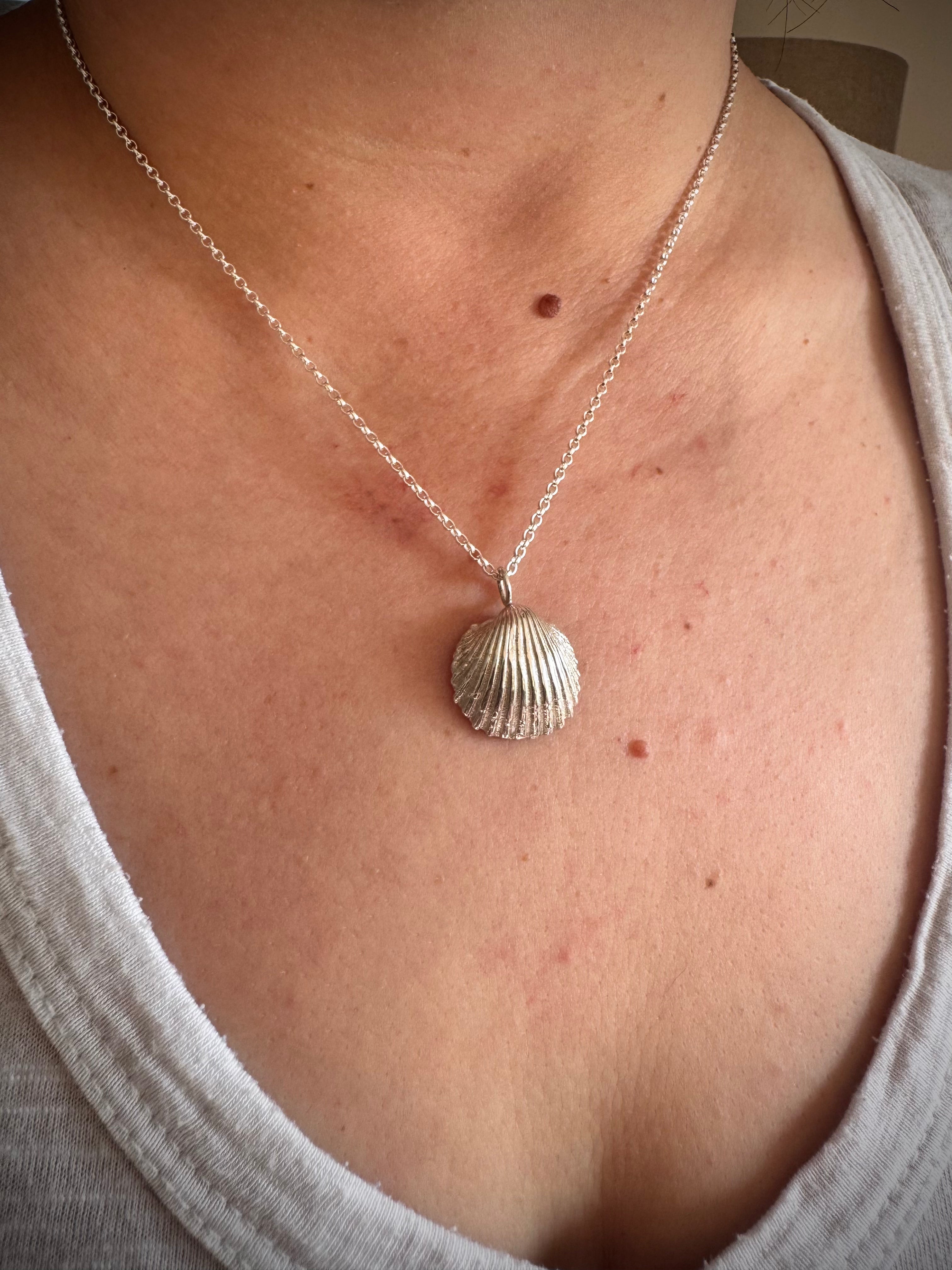 A silver shell pendant necklace worn by a person, shown close-up against their chest and neck. The necklace features a textured, ribbed seashell pendant hanging from a delicate silver chain.