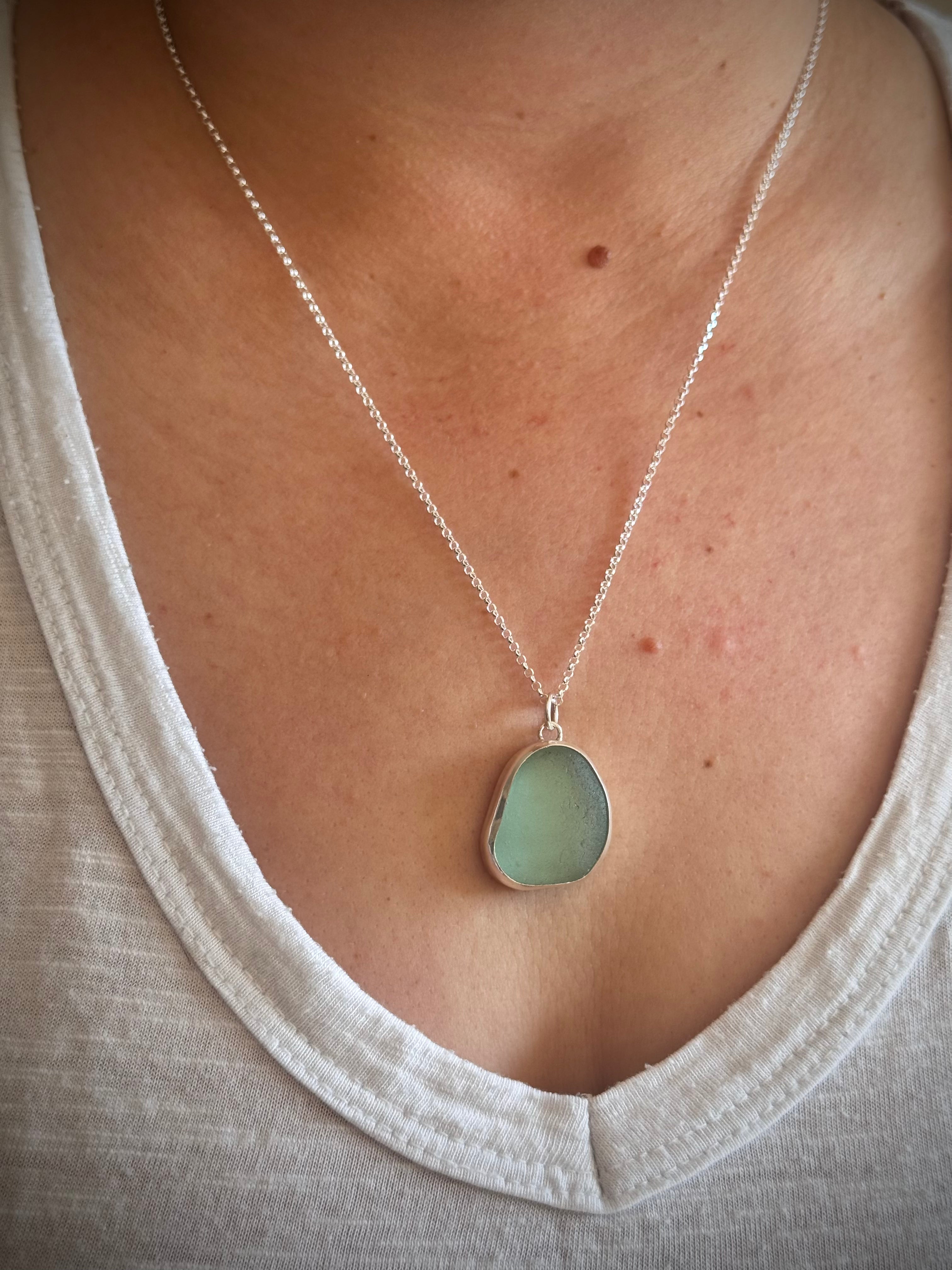 A close-up of a person wearing a silver chain necklace with a flat, irregular light aqua green sea glass pendant, styled with a white top.